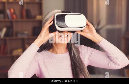 Smiling Elderly Caucasian Woman Using Virtual Reality Headset At Home, Technology Concept Stock Photo