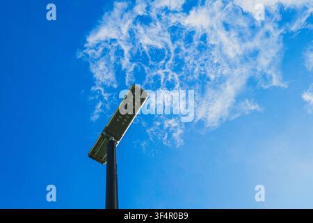 A low-angle shot of a modern outdoor LED solar street lamp on a metal pole against a bright blue sky with wispy white clouds. Stock Photo