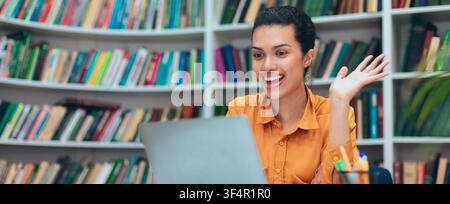 Smiling Young Hispanic Woman Using Laptop In Library, Engaged In Online Learning Stock Photo