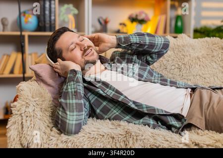 Young man on home sofa meditates with wireless headphones and slow breathing while listening to calm music. Adult guy on home sofa restores emotional balance and relaxes deeply in quiet home comfort. Stock Photo