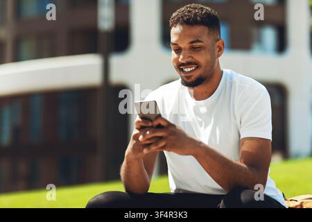 Smiling Black Man Using Smartphone Outdoors On Grass, Happy Lifestyle Concept Stock Photo