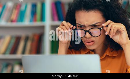 Worried Young Woman Analyzing Data On Laptop In Library, Focused Expression Stock Photo