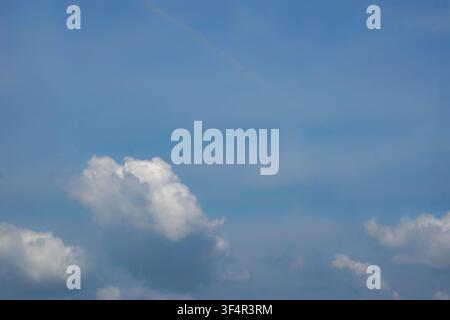 White cumulus clouds float across a bright blue sky with soft gradients and a faint contrail above the horizon. Stock Photo