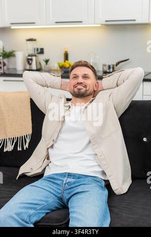 Young man at home leans into backrest places hands behind head and releases tension with slow breath. Caucasian guy on sofa stretches lightly closes eyes and shows calm relief comfort and gentle rest. Stock Photo