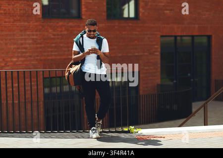 Young Black Man Texting On Phone While Leaning Against Railing, Outdoor Setting Stock Photo
