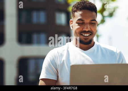 Smiling Young Black Man Working On Laptop Outdoors, Focused On Task, Business Concept Stock Photo