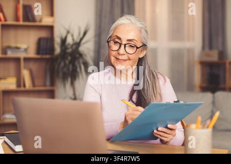 Senior Woman Writing Notes At Home Office While Using Laptop, Business Concept Stock Photo