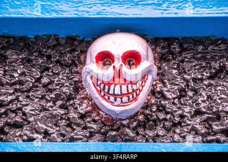 A scary skull mask with a smile and red eyes, set against a background of dark stones and a blue surface, on a Kora pagoda in Dharamsala, Himachal Pra Stock Photo