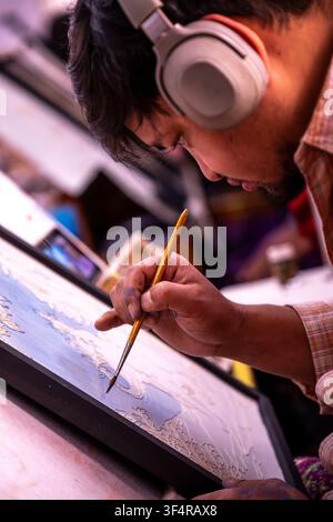 Norbulingka Institute, Sidhpur, Himachal Pradesh, India. An artist, deeply focused, uses a fine brush to paint a detail on a work of art. He is wearin Stock Photo