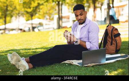 Focused Black Male Student Working On Laptop Taking Notes In Park, Education Concept Stock Photo