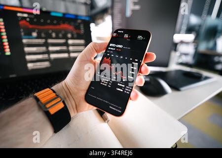 Bitcoin stock market crash phone hand financial chart office. Male hand holding smartphone showing red cryptocurrency falling chart on screen inside m Stock Photo