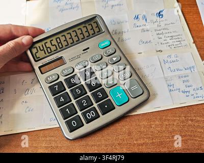 A hand holds a calculator showing numbers while papers with budget notes are spread on a table, indicating expense tracking at home. Stock Photo