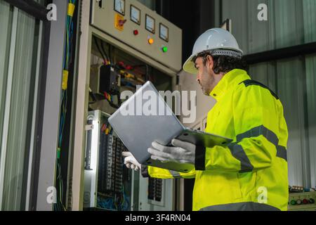 Male electrical engineer holding laptop inspecting industrial electrical cabinet storehouse. Concept of heavy machinery power supply, electrical maint Stock Photo