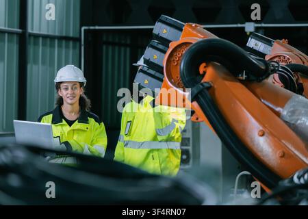 Automation technician woman holding laptop looking camera while inspecting orange robotic arm plant. Concept of industrial workforce team, artificial Stock Photo