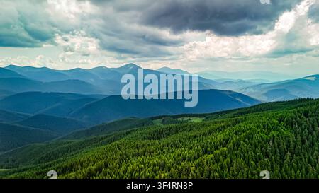 The view opens to a countryside beyond green ridges. Patches of sun drift across meadows and dark woods as layers of mountains step into the distance Stock Photo