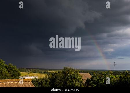 Brooding storm clouds sweep over open countryside as a delicate rainbow appears in the brightening gap above sunlit trees and roofs the dramatic sky c Stock Photo