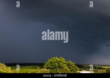 A massive wall of storm cloud blankets the horizon as vivid greens glow under stray sunlight the countryside bracing for rainfall with a powerful cont Stock Photo