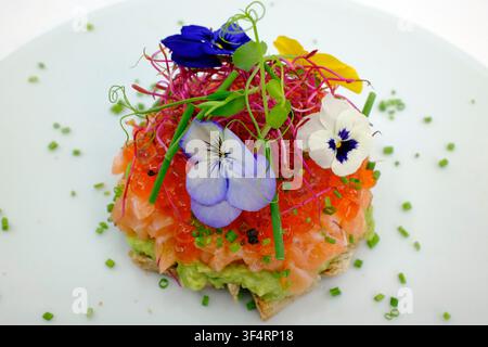 A gourmet dish of salmon tartare and ikura on avocado, garnished with microgreens, chives, and edible flowers on a white plate. A gourmet Asian-style Stock Photo