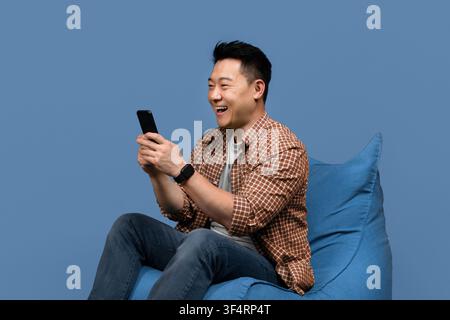 Smiling Asian Man Using Smartphone While Sitting On Bean Bag Chair, Relaxed Atmosphere Stock Photo