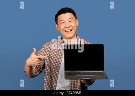 Smiling Asian Male Holding Laptop With Blank Screen, Blue Background, Mockup Stock Photo