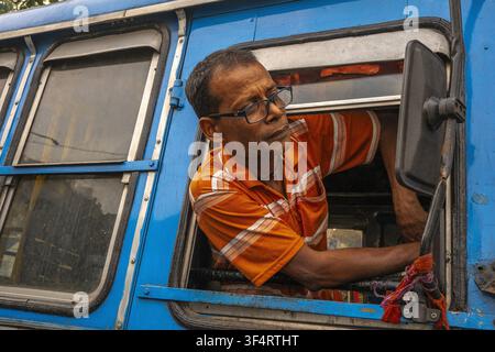 A bus driver wearing glasses and an orange-striped shirt looks out the window of a blue bus in Kolkata, West Bengal, India Stock Photo