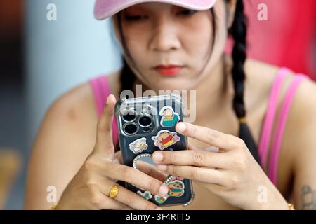 A young woman with braided hair is using her smartphone, which is decorated with stickers of cartoon characters. Social media. Pattaya, Thailand. Stock Photo