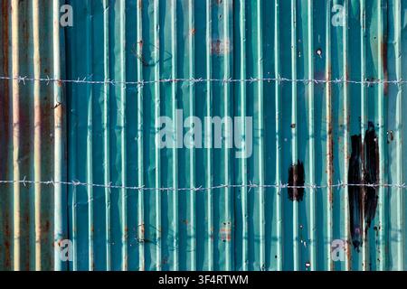 A corrugated metal fence, partially rusted and painted green, is crisscrossed by barbed wire. The image highlights the texture and the shadows created Stock Photo