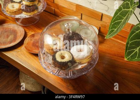 Selection of assorted gourmet donuts displayed under a clear cake dome on a wooden counter inside a modern bakery and cafe. Stock Photo