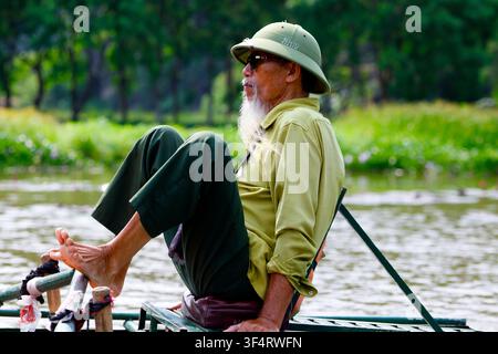 A middle-aged man, wearing a bodo helmet and sunglasses, is rowing a traditional boat. He uses his feet to maneuver the oars. Tam Coc, Vietnam. Stock Photo