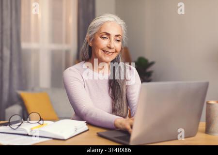 Happy Elderly Caucasian Woman Using Laptop At Home, Engaged In Work Or Communication Stock Photo