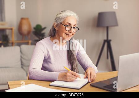 Smiling Elderly Caucasian Woman Working At Home Office, Writing In Notebook, Business Concept Stock Photo