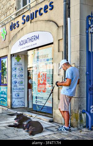 Casually dressed man leaning on wall browsing smartphone whilst holding resting dog on leash - La Roche Posay, Vienne (86), France. Stock Photo