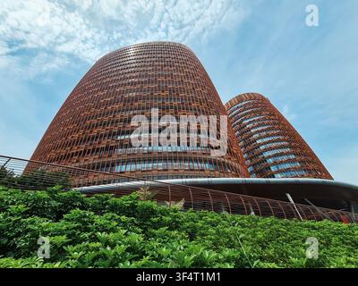 Low angle shot of twin cylindrical corporate buildings featuring unique reddish textured facades and lush garden borders against a bright sky. Stock Photo