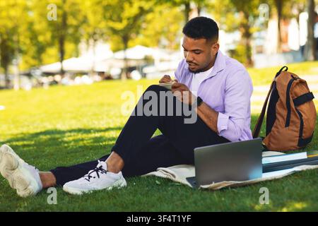 Focused Black Male Student Writing Notes In Park, Using Laptop For Study, Educational Concept Stock Photo