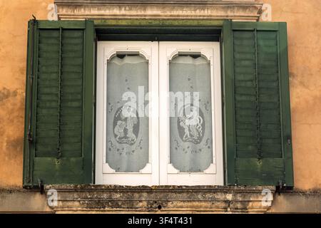Classic window with open green shutters & delicate crochet patterned window covering on an orange stucco facade, Ragusa Ibla,  Sicily, Italy Stock Photo