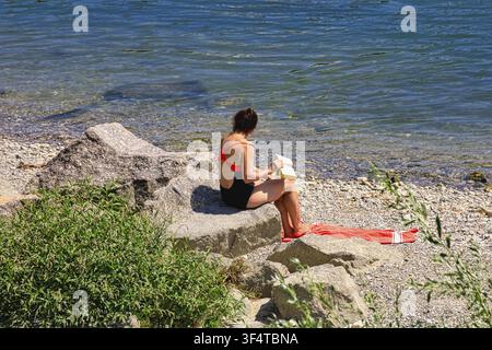 A woman sitting on stones on the shore and reading a book, Urban Bathing, Rhine, Leisure Activity, Bathing, Relaxing, Enjoying the Sun, Rheinbord Base Stock Photo