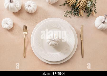 Minimal autumn table setting with white ceramic plates, decorative pumpkins, dried grasses and golden cutlery on beige background. Flat lay with copy Stock Photo