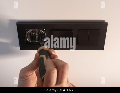 Hand holds a screwdriver to tighten a black electrical socket on a white wall. The person performs a home repair task fixing the socket panel in place Stock Photo