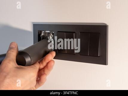 Hand installs a black electrical socket into a dark frame on a white wall. The electrician fits the socket mechanism into the wall panel during a home Stock Photo