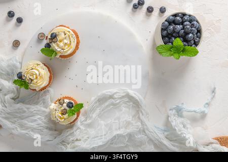 An overhead flat lay composition features three cupcakes with white frosting and a bowl of blueberries on a textured white surface. Stock Photo