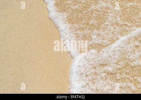 Soft ocean wave foam gently washing over fine golden sand on a tropical beach, high angle top-down view showing natural coastal texture and copy space Stock Photo