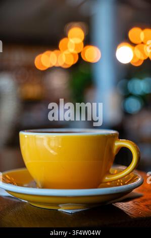 A yellow coffee cup sits on a white saucer at a table inside a cafe with blurred lights. Stock Photo