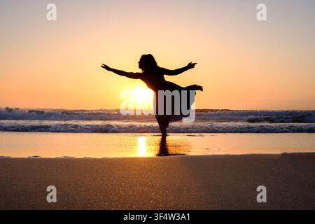 Woman silhouetted against a vibrant sunrise, balancing on one leg with outstretched arms on the beach Stock Photo