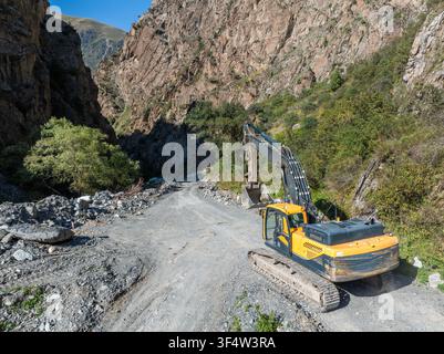 Excavator rests on a rough, unpaved road winding through a mountainous gorge. Steep rocky slopes rise dramatically on either side, showcasing a rugged Stock Photo