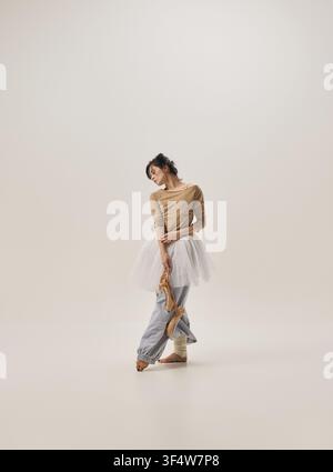 Young ballerina standing in relaxed crossed leg pose wearing tutu and warmup pants on neutral background Stock Photo