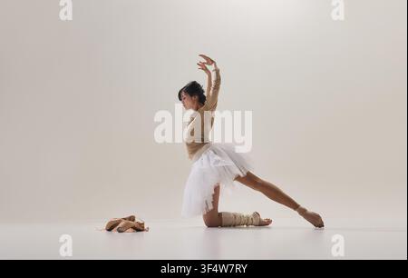 Ballerina performing kneeling side stretch beside ballet shoes on white studio background Stock Photo