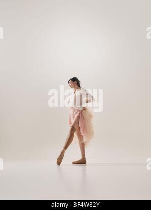 Ballerina in flowing pink dress walking thoughtfully across minimalist white studio Stock Photo