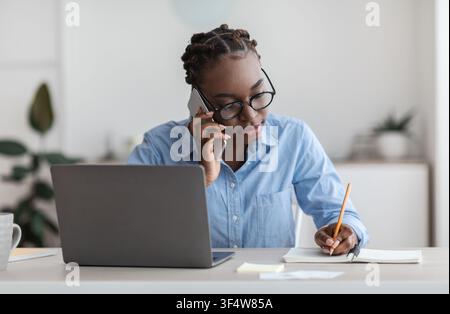 Black Female Office Worker Talking On Cellphone With Client And Taking Notes Stock Photo