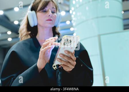 A woman wears headphones while interacting with her smartphone in a public space, focusing on the screen with her finger Stock Photo