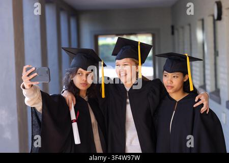 Diverse female graduates taking selfie with phone in corridor, wearing gowns, caps, holding diploma Stock Photo